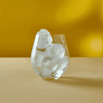 A glass with large ice cubes and Mother Root Ginger sits on a beige surface with a yellow background.