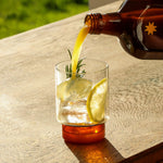 Mother Root Ginger by Mother Root is poured from a brown bottle into a glass of lemon, ice, rosemary, and blossom honey.