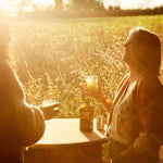 Two women laugh over drinks of Mother Root Ginger by Mother Root at a sunlit outdoor table.