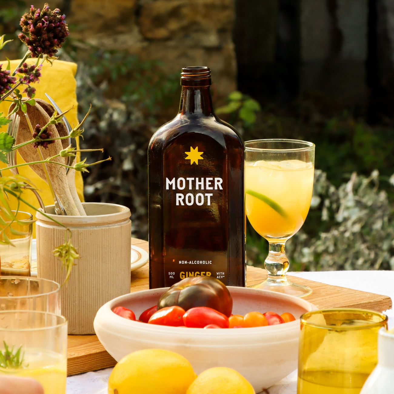 A bottle of Mother Root Ginger by Mother Root on a table with apple cider vinegar, lemons, and a glass of yellow drink.