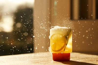 A fizzy drink with lemon slices splashes on a sunny table, sparkling droplets caught in midair.
