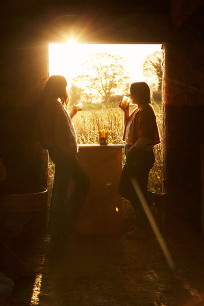 Two people stand in a doorway at sunset, chatting and holding drinks as golden sunlight streams in behind them.