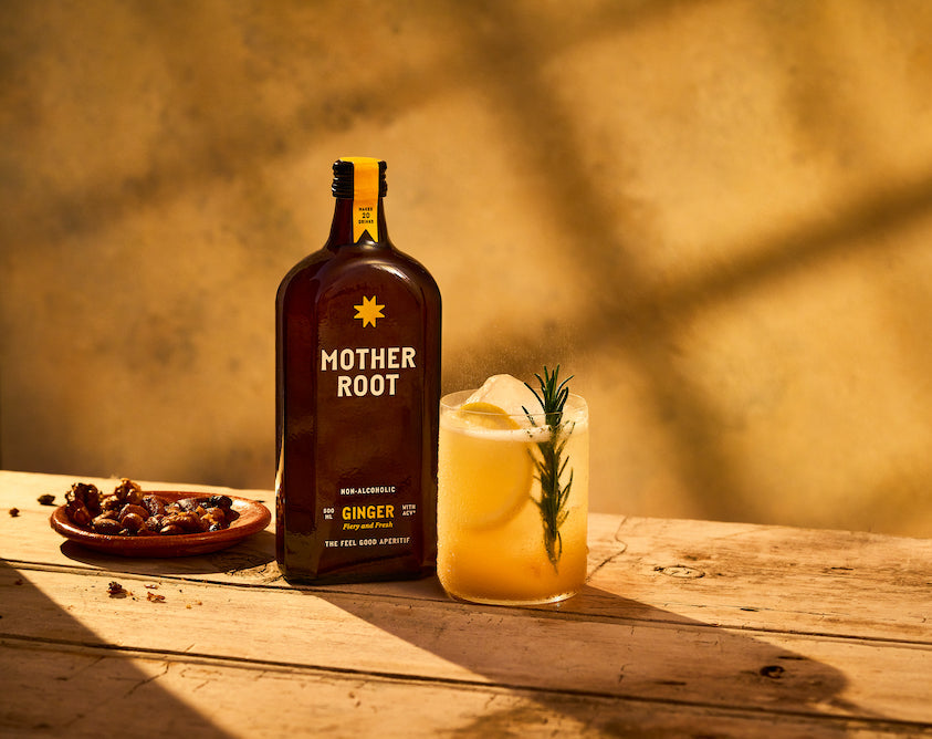 A bottle of Mother Root ginger, a drink with ice and rosemary, and a small dish of nuts on a wooden table.