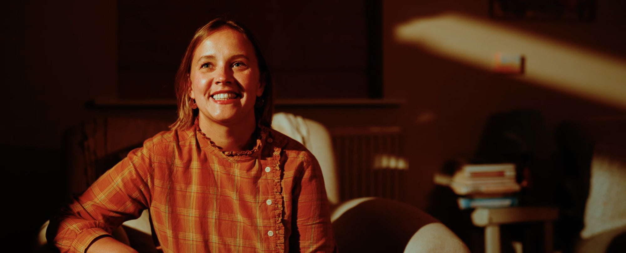 A smiling person in an orange plaid shirt sits indoors, warmly lit by sunlight through a window, creating a cozy atmosphere.