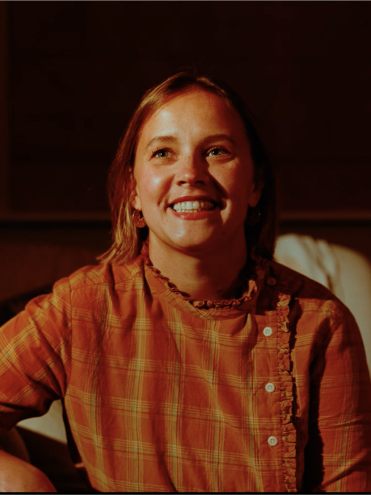 Smiling woman in an orange plaid shirt sits indoors in warm lighting, enjoying a cozy, inviting atmosphere.