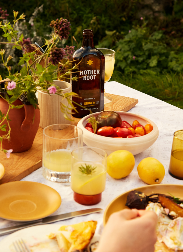 Outdoor table with Mother Root ginger drink, glasses, lemons, tomatoes, flowers, and a plate of food on a sunny afternoon.