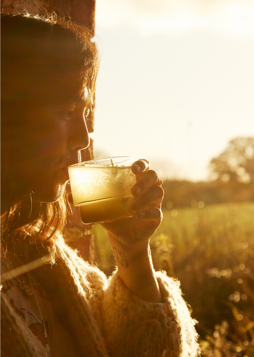 A person with long hair enjoys a refreshing drink outdoors in warm, golden sunlight.