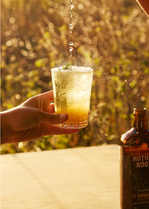 A hand holds a fizzy drink with herbs as liquid is poured in, outdoors in warm sunlight. A bottle sits on the table.