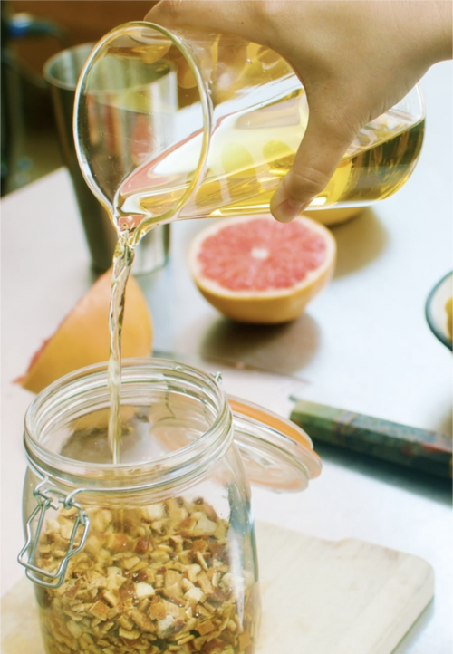 A hand pours liquid from a glass pitcher into a jar with dried fruit; fresh grapefruit sits in the background.