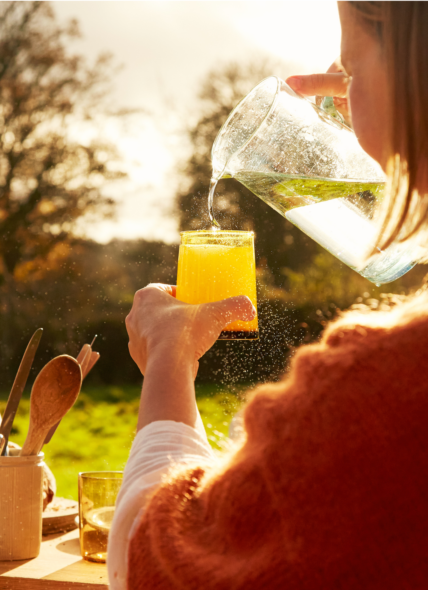Someone pours juice from a glass pitcher into a glass outdoors in warm, golden morning sunlight.