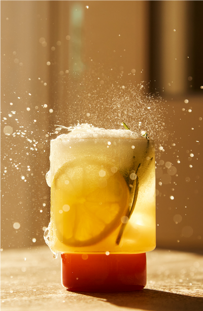 A fizzy spritzer with lemon slices and rosemary in a glass, bursting with bubbles and splashes in warm sunlight.