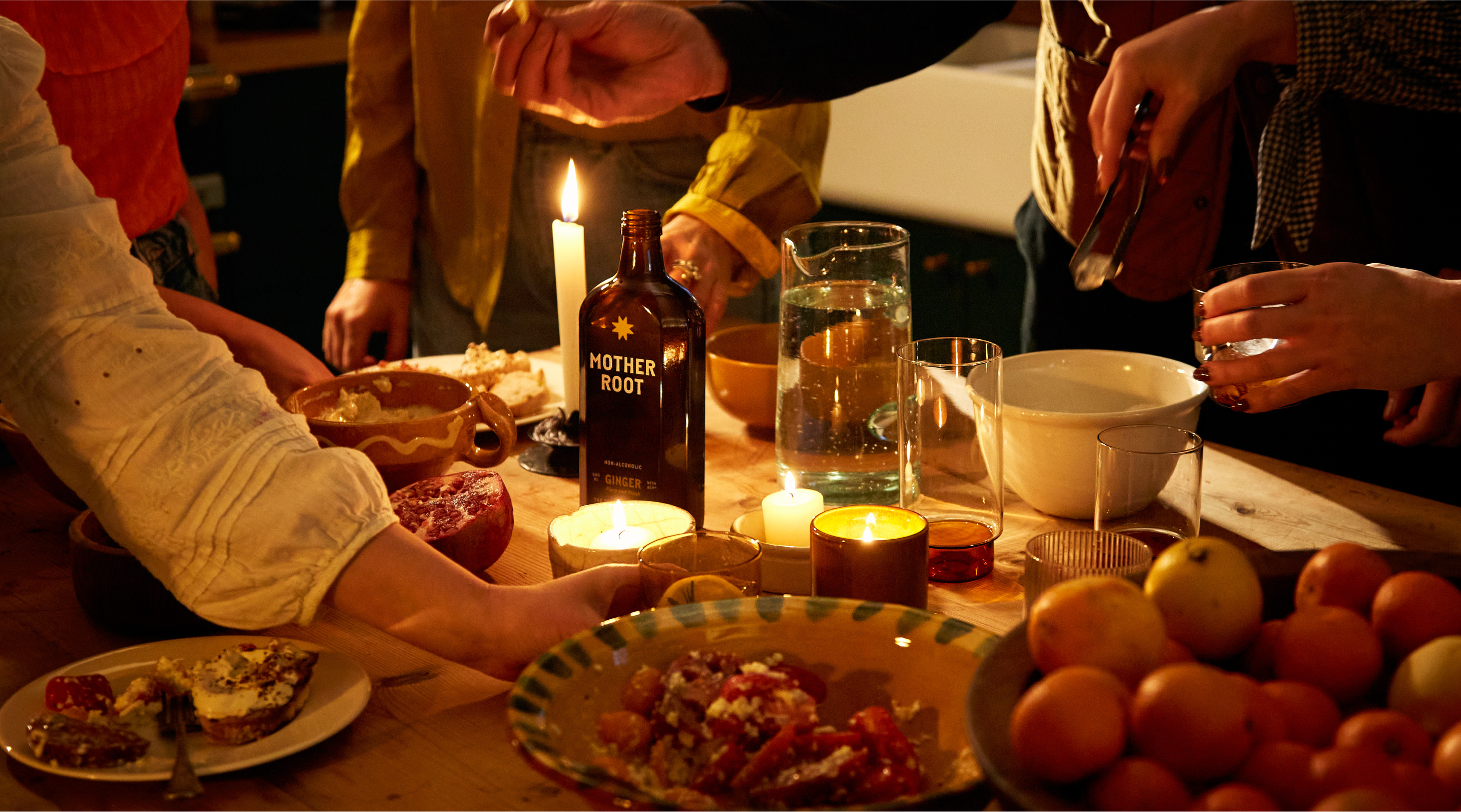 People gathered around a candlelit table with food, drinks, and fresh fruit, reaching for various dishes.