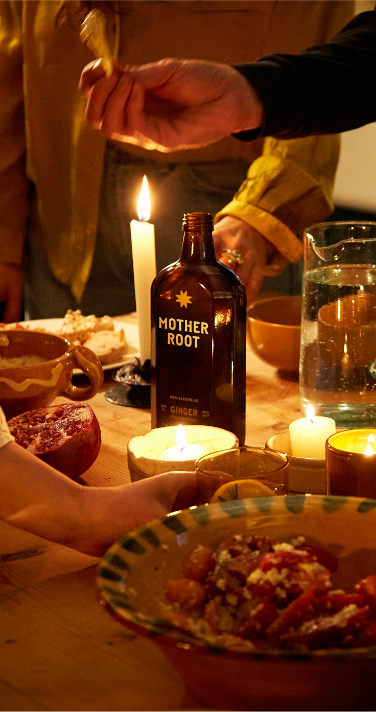 A bottle of Mother Root ginger drink sits on a candlelit dinner table as friends reach for delicious food.