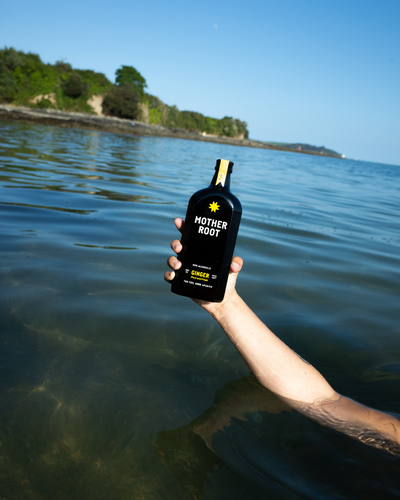 A hand holds a Mother Root Ginger bottle above the water with trees and blue sky in the background.