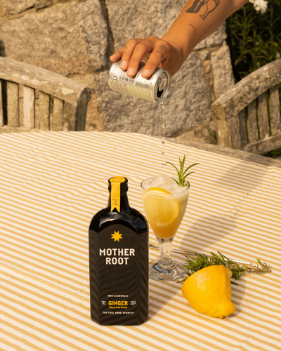 A hand pours soda into a glass with ice and lemon beside a Mother Root bottle on a striped tablecloth.