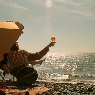 Person relaxing on a beach chair under a yellow umbrella, holding up a glass as the ocean glistens in the background.