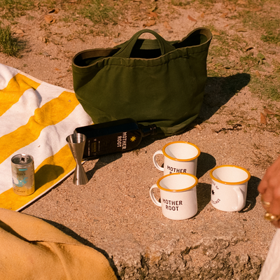 Picnic setup on sandy ground with three mugs, a green bag, a bottle, can, jigger, and towel.